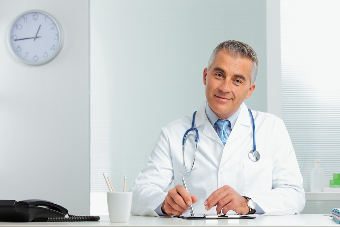 A male doctor is sitting at his desk, wearing a white coat, discussing using Ace QBank to prepare for the MCCQE1 exam.