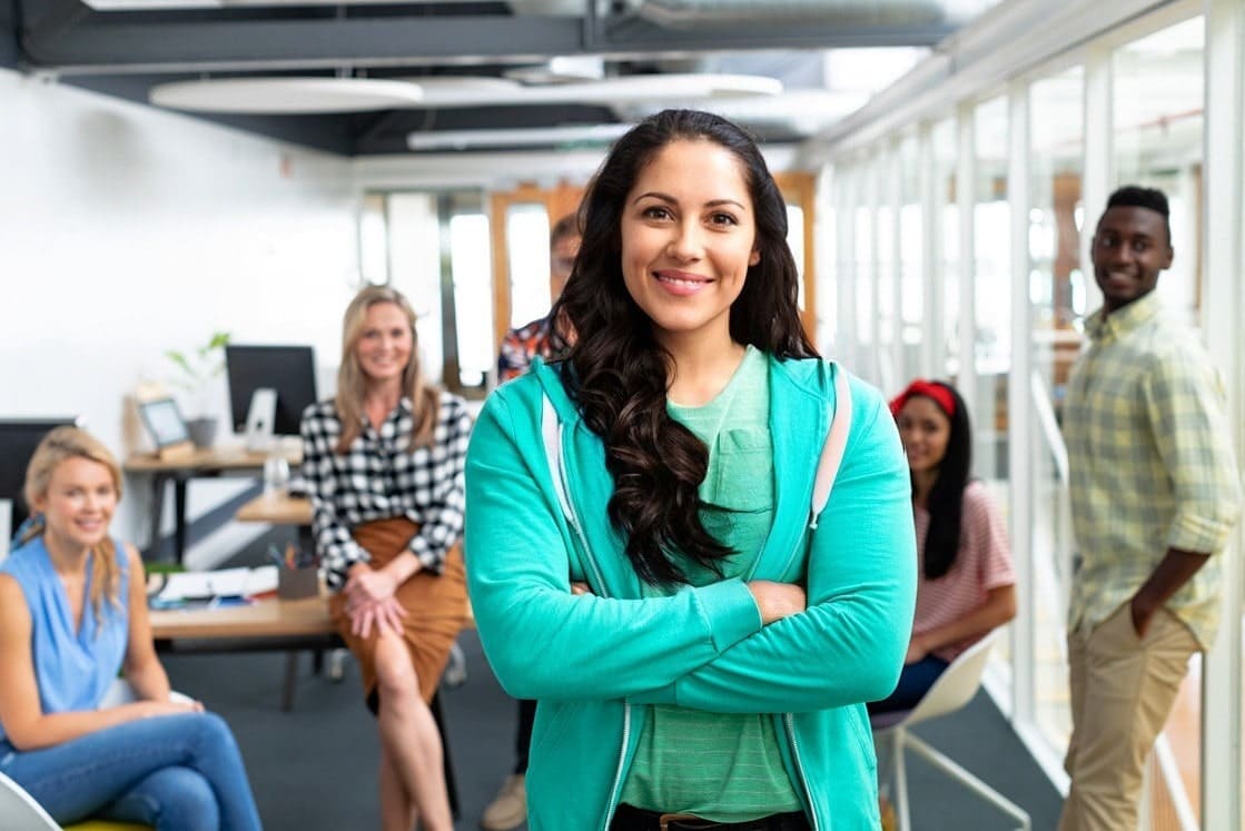 A confident doctor stands with arms crossed in a modern office environment with colleagues in the background, preparing for PLAB from Ace QBank.