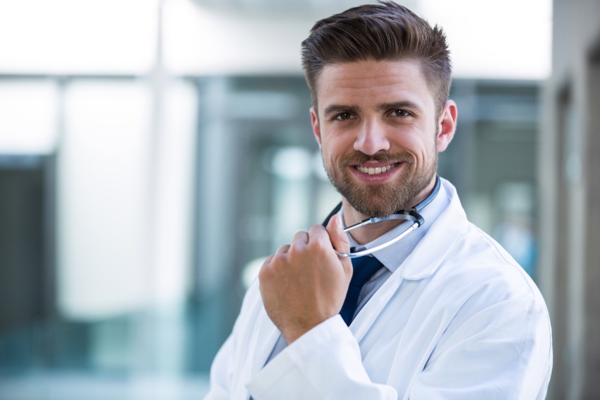 A male doctor with a beard and a smiling expression wears a white coat and holds a stethoscope around his neck in an indoor setting, exuding confidence as he prepares for the MCCQE1 exam with Ace QBank.