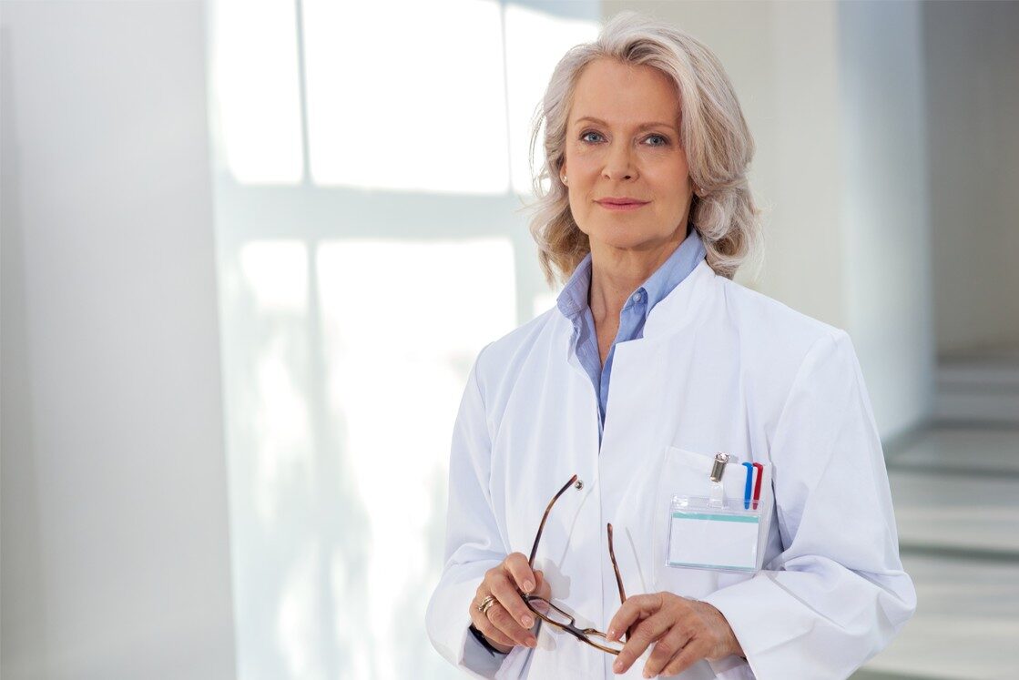 An older woman with gray hair, wearing a white lab coat and holding eyeglasses, stands in a well-lit room. She has a name tag and pens in her coat pocket, indicating her the Role of MCCQE1 in Doctor Salaries.