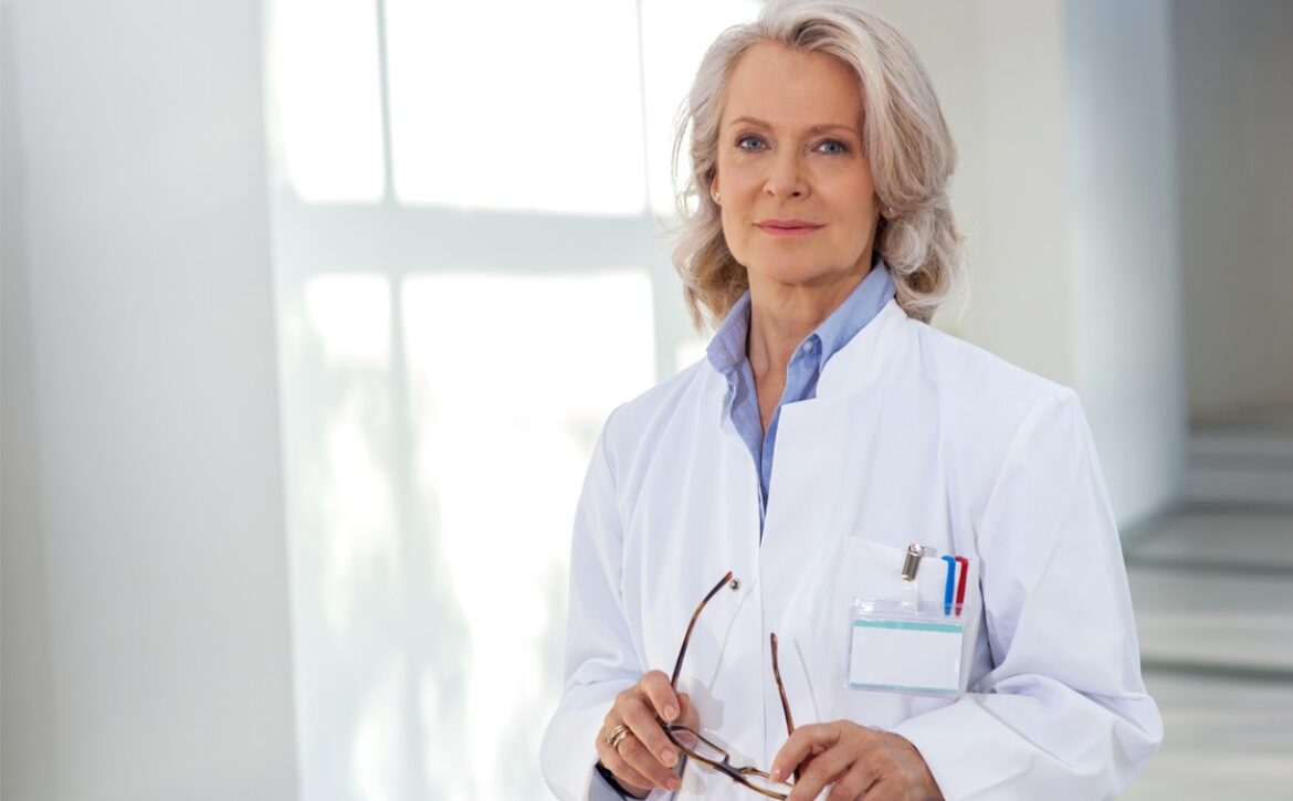 An older woman with gray hair, wearing a white lab coat and holding eyeglasses, stands in a well-lit room. She has a name tag and pens in her coat pocket, indicating her the Role of MCCQE1 in Doctor Salaries.