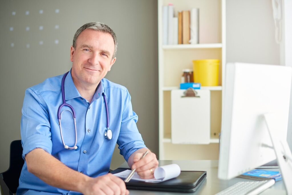 A doctor in a blue shirt with a stethoscope around his neck sits at a desk, smiling, with a pen in hand. A computer monitor and bookshelves are visible in the background, perhaps filled with resources like Ace QBank for those wanting to know how to overcome MCCQE1 exam anxiety.