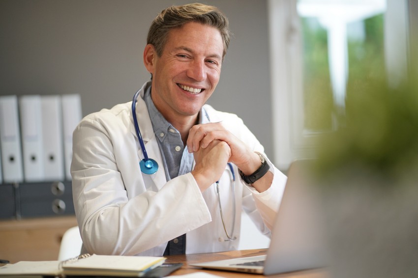 A smiling doctor wearing a white coat and stethoscope sits at a desk with folders and a laptop in an office, reflecting on how the role of MCCQE1 in doctor salaries impacts his career.