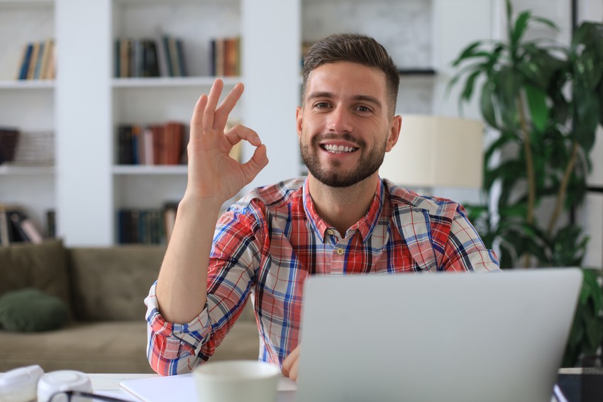 A man in a plaid shirt sits at a desk in a well-lit room with bookshelves, smiling and making an "OK" gesture with his hand while looking at the camera, as if to suggest you try Ace QBank demo questions.