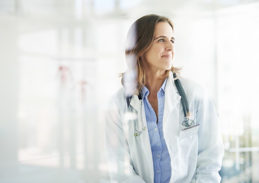 A woman with long hair, wearing a white lab coat and a stethoscope around her neck, looks to the side in a bright, modern medical facility, clearly pondering which specialty to choose.