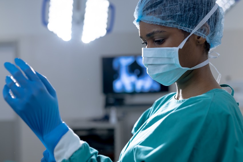A female surgeon in scrubs puts on blue surgical gloves in an operating room, with screens in the background encouraging women to break the glass ceiling with MCCQE1.