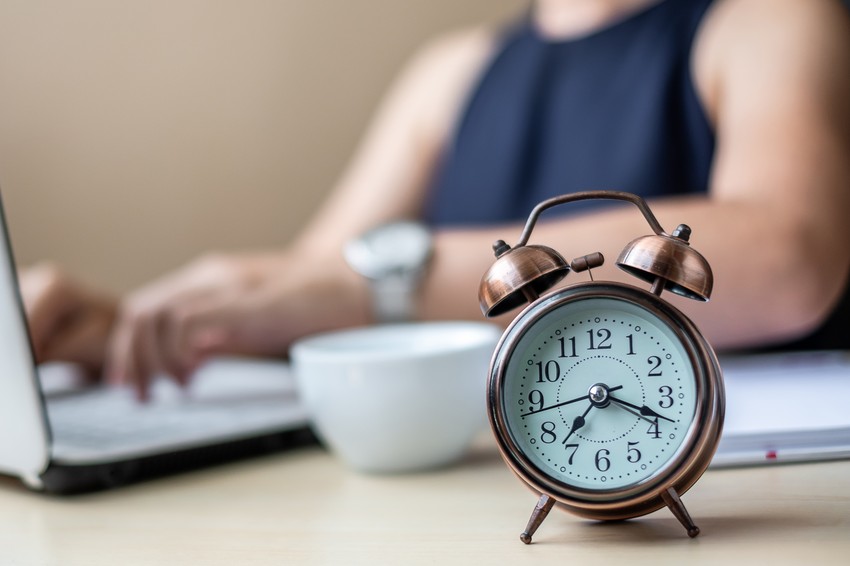 A vintage copper alarm clock on a desk, showing 10:10, with a blurred background of a person studying on a laptop for MCCQE1 with Ace QBank.