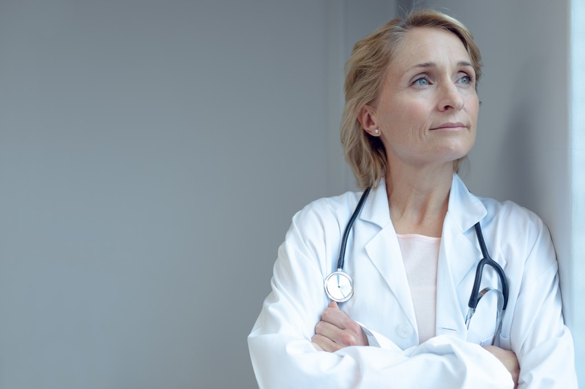 A middle-aged female doctor in a white coat with a stethoscope, looking thoughtful and gazing upwards, thinking about encouraging women to break the glass ceiling with MCCQE1.