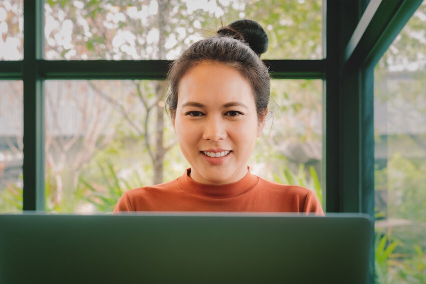 A person with a bun hairstyle, wearing an orange top, is smiling while looking at a laptop. While studying for the MCCQE1 exam preparation with Ace QBank.