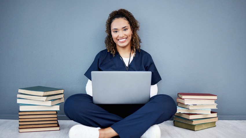 A person in scrubs sits on the floor against a gray wall, smiling and working on a laptop. They're surrounded by stacks of books, clearly deep into their MCCQE1 exam preparation.