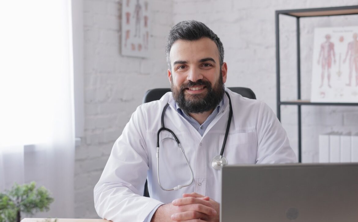 A bearded man in a white coat with a stethoscope around his neck sits at a desk and smiles at the camera. Medical charts, MCC objectives, and Ace QBank materials are visible on the wall in the background.