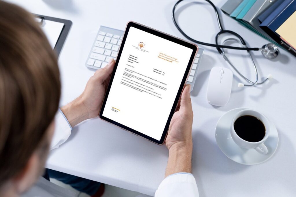 A person is holding a tablet displaying their MCCQE1 Result. A keyboard, mouse, stethoscope, coffee cup, and files are neatly arranged on the white desk.