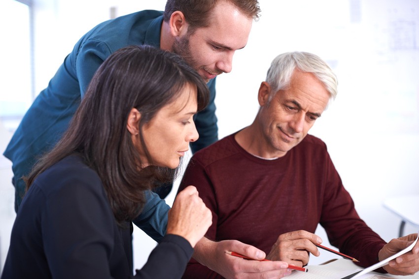 Three people are engaged in a discussion, looking at documents. Two are seated while one stands, leaning in to examine the papers closely. They seem focused and intent, likely deep into MCCQE1 exam preparation as part of their group study session.