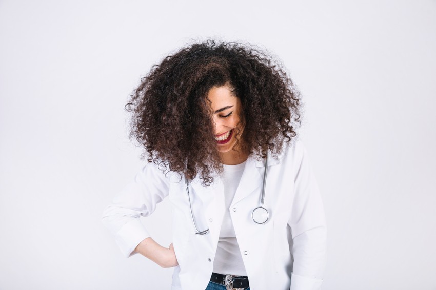 A smiling person with curly hair, wearing a white coat and stethoscope, stands against a plain background, ready to conquer the future with a high score of MCCQE1 with Ace QBank aid.