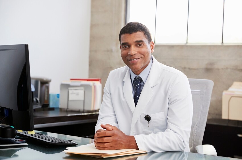 A person wearing a white lab coat and a tie sits at a desk with a computer monitor and files, smiling and looking at the camera, feeling confident about their MCCQE1 result thanks to Ace QBank.