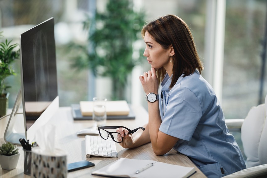 A woman in medical scrubs sits at a desk with a computer, holding eyeglasses in one hand and resting her chin on the other, looking focused as she reviews her MCCQE1 Result.