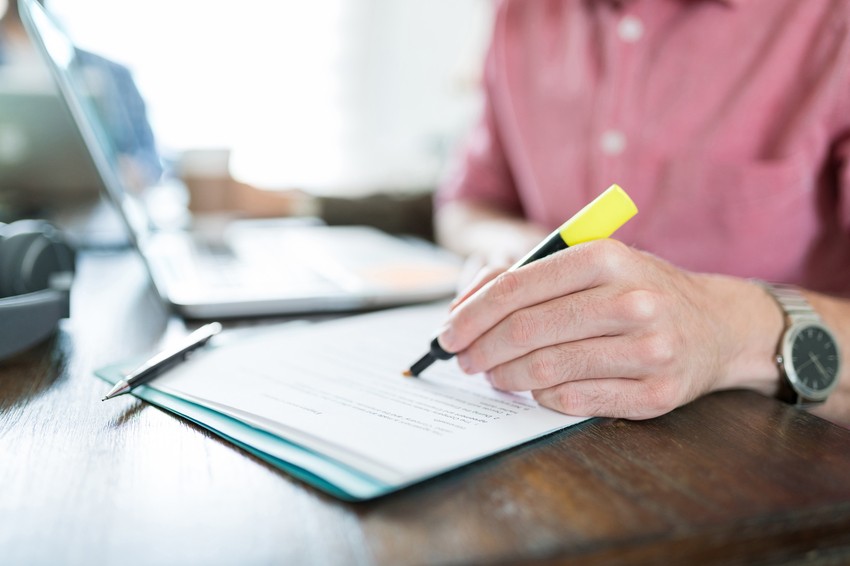 A person in a red shirt is highlighting text on a document with a yellow highlighter next to a laptop on a wooden desk, diligently focusing on MCC objectives for MCCQE1 exam preparation.