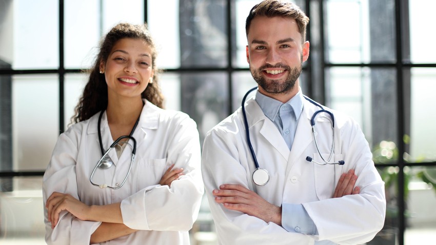 Two doctors, wearing white coats and stethoscopes, stand with their arms crossed, smiling in front of a windowed background. Their confident posture reflects their mastery of MCC objectives and expertise honed with Ace QBank.