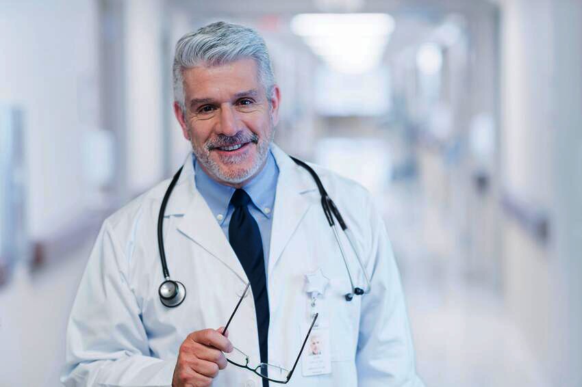 A middle-aged male doctor with gray hair, wearing a white coat and stethoscope, smiles confidently while holding eyeglasses in a bright hospital corridor, having just reviewed the latest MCCQE1 Qbank update.