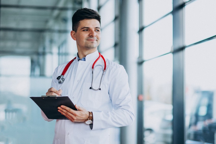 A male doctor in a white coat stands by a large window, holding a clipboard and pen. He has a stethoscope around his neck and is looking to the side with a slight smile, perhaps contemplating the Ace QBank online question bank for MCCQE Part I preparation.