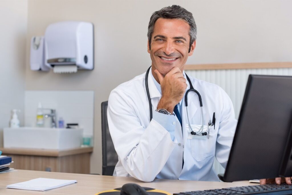 A doctor in a white coat and stethoscope sits at a desk, smiling, with a hand on his chin. In the background, there is a wall-mounted paper towel dispenser and shelves with medical supplies for MCCQE1 exam preparation.