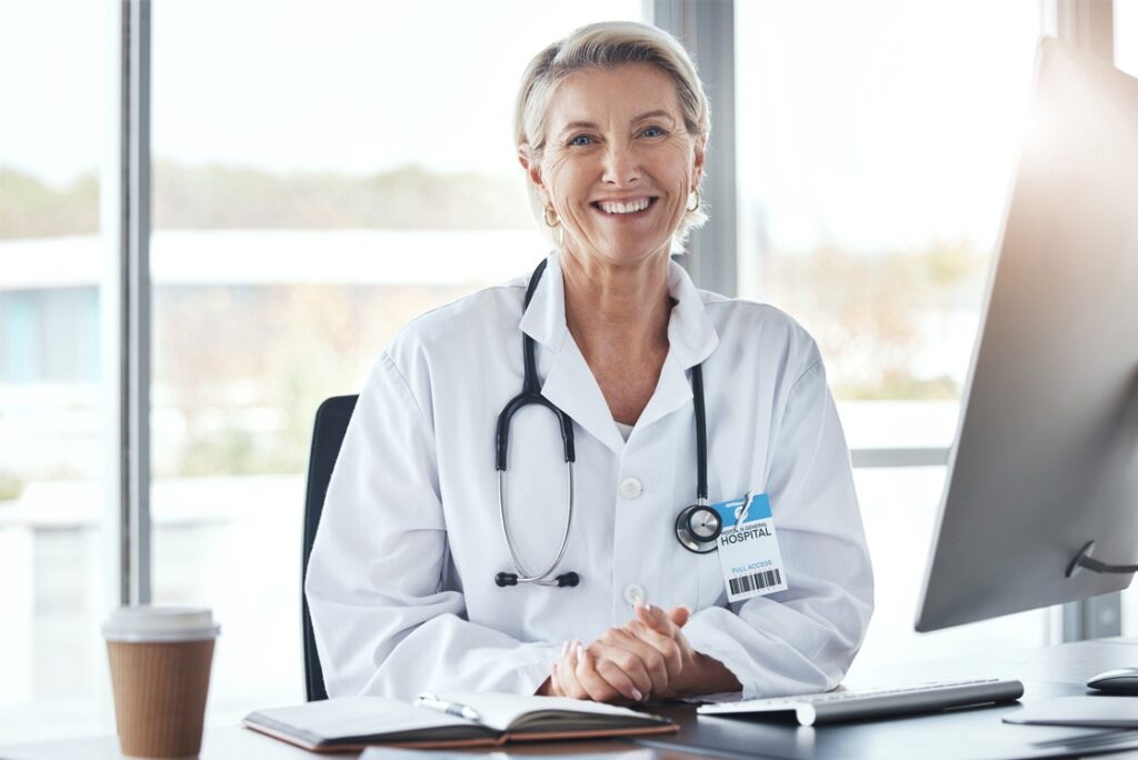 A smiling doctor in a white coat with a stethoscope around their neck sits at a desk with a computer, notebook, and coffee cup, diligently working on MCCQE1 exam preparation.