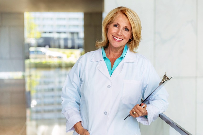 A middle-aged woman in a white lab coat holds a clipboard and smiles while standing in a modern office, embodying focus and discipline essential for MCCQE1 exam preparation.