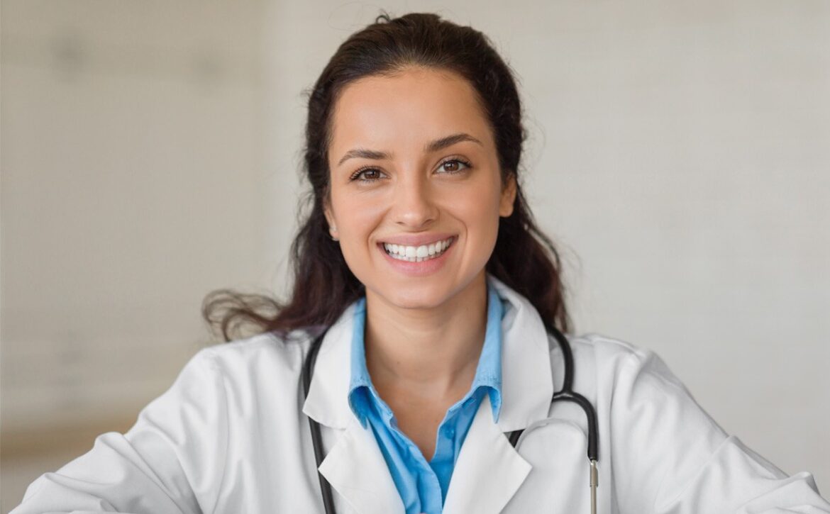 A woman in a white medical coat and blue shirt, wearing a stethoscope, smiles at the camera. She is seated in a neutral indoor setting, effortlessly balancing health and study as she prepares for the MCCQE1 exam with Ace QBank.