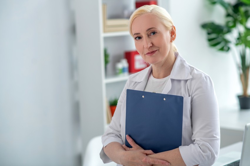 A woman with blonde hair, dressed in a white coat, is holding a blue folder. She stands indoors with a neutral expression, with shelves and plants in the background, possibly taking a break from MCCQE1 exam preparation to avoid burnout.