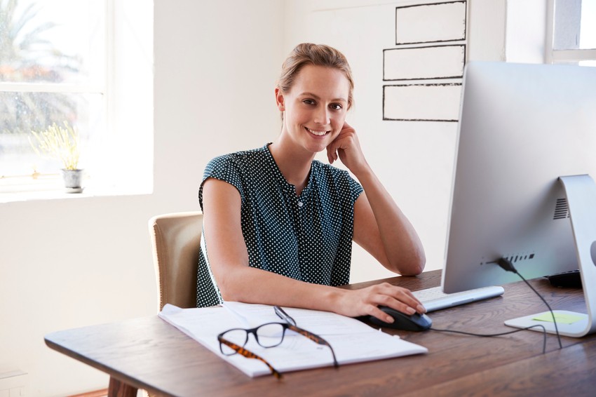 A woman sits at a desk, smiling, with her hand on a computer mouse. Papers and eyeglasses are on the desk in front of her, alongside materials for MCCQE1 exam preparation, and there is a window in the background.