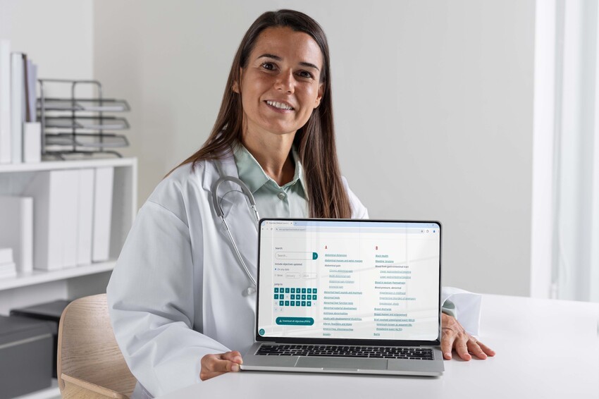 A doctor in a white coat holds a laptop displaying a medical interface, seated at a desk in a modern, well-lit office. The screen shows resources for MCCQE1 exam preparation, aligning with MCC objectives to ensure top-notch proficiency.