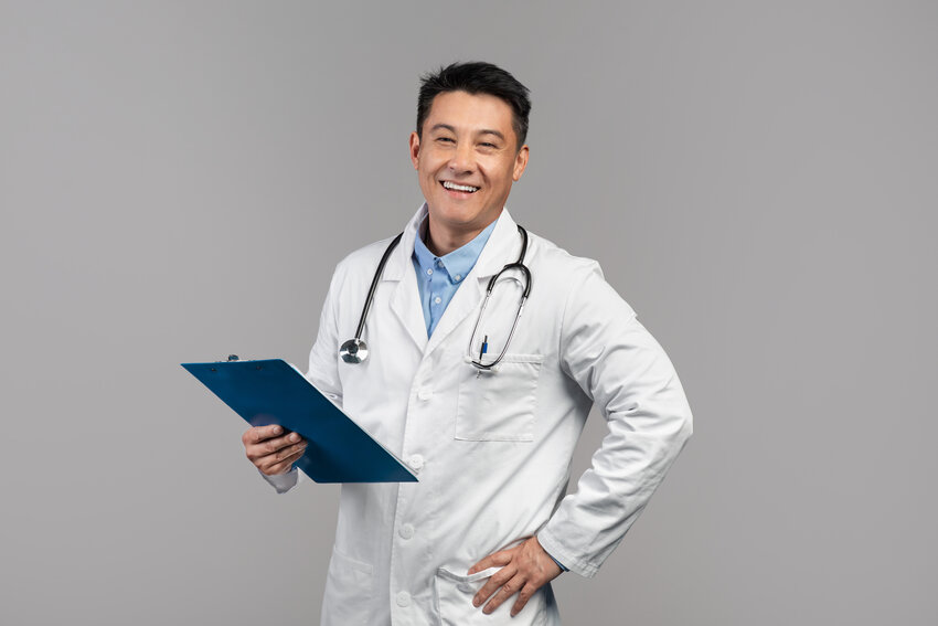 A man in a white medical coat and blue shirt, wearing a stethoscope, smiles at the camera. He is seated in a neutral indoor setting, effortlessly balancing health and study as she prepares for the MCCQE1 exam with Ace QBank.