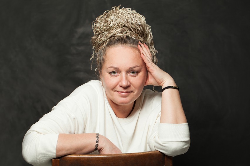 Woman with light-colored braided hairstyle and white sweater leans on a wooden chair, resting her head on her hand, as she contemplates Ace QBank for MCCQE1 exam preparation in front of a dark background.