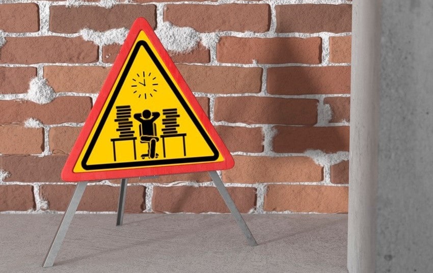 Triangular caution sign depicts a person overwhelmed by stacks of paperwork at a desk, placed against a brick wall background. Avoid procrastination to boost MCCQE1 prep with Ace QBank!