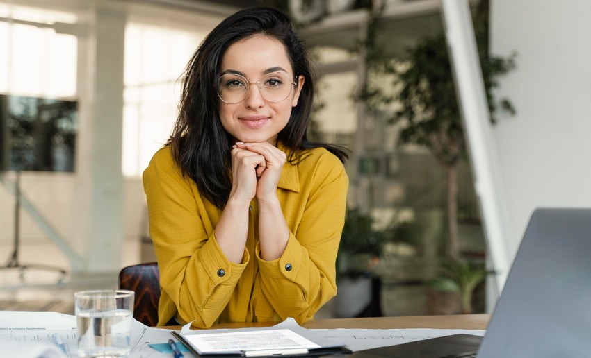 A person with glasses and a yellow shirt sits at a table, hands under chin, surrounded by papers, a glass of water, and a laptop. They're engrossed in MCCQE1 exam preparation using Ace QBank. An indoor environment with plants is in the background.