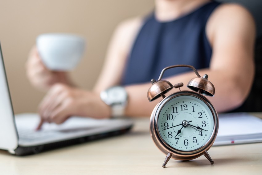 An alarm clock sits on a desk in the foreground, with a person using a laptop and holding a cup in the blurred background. The visible Ace QBank logo hints at their preparation for the MCCQE1, battling procrastination one study session at a time.