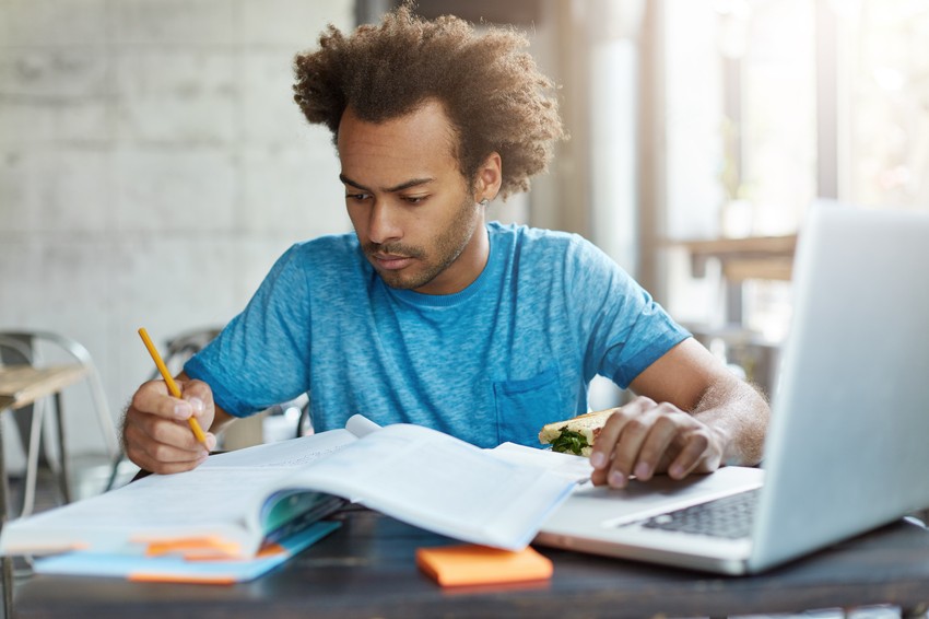 A person in a blue shirt is writing in a notebook while holding a sandwich and using a laptop at a table with an open book and sticky notes, balancing health and study as they focus on their MCCQE1 exam preparation.