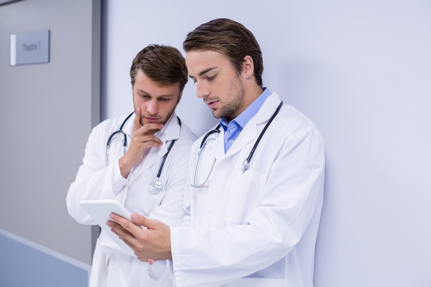 Two male doctors in white lab coats and stethoscopes stand in a hospital corridor. One holds a tablet while both look at it intently, likely reviewing MCCQE1 prep materials with Ace QBank.