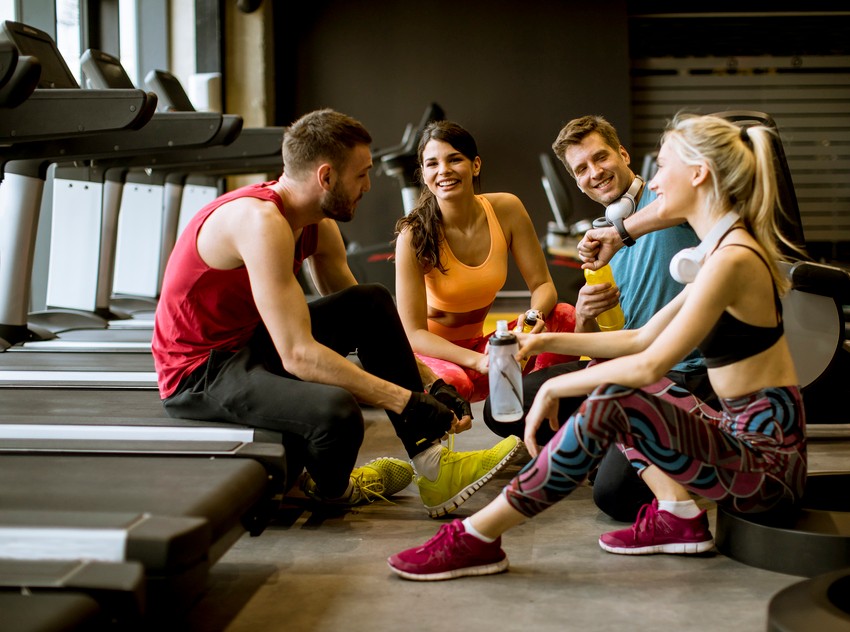 Four people in athletic wear sit on the gym floor, laughing and taking a break near treadmills. They chat about balancing health and study, particularly how Ace QBank helped one of them ace their MCCQE1 exam.