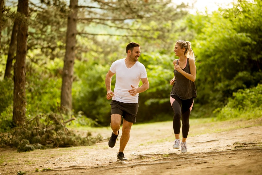 Two people are jogging on a dirt trail in a forested area, smiling at each other. Trees and greenery surround them, as they discuss how balancing health and study is key to their success with MCCQE1 exam preparation.