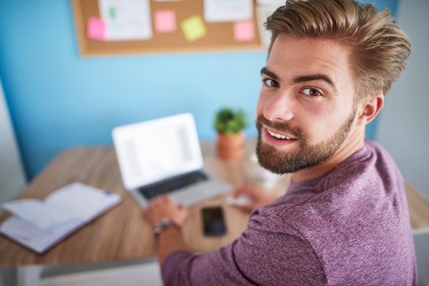 A man with a beard and short hair is sitting at a desk, looking up at the camera. He is using a laptop for his MCCQE1 exam preparation, surrounded by a notebook, smartphone, and potted plant. A bulletin board is in the background.