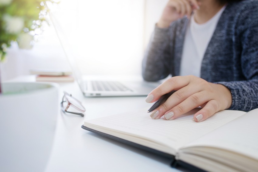 A person sits at a desk with a laptop, holding a pen and writing in a notebook. Glasses are placed on the desk nearby, hinting at moments of procrastination while preparing for the MCCQE1 exam with Ace QBank.