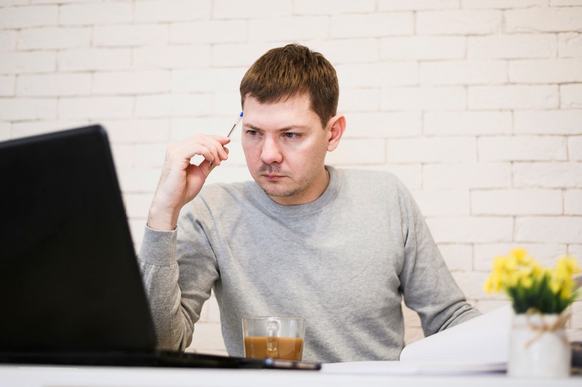 A person in a gray sweater holding a pen and looking at a laptop screen with a serious expression, likely deep into their MCCQE1 exam preparation using Ace QBank. There's a coffee cup on the table and a small plant in the background, hinting at efforts to combat procrastination.