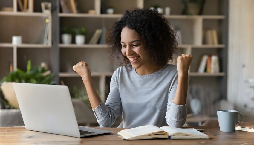 A woman with curly hair, wearing a gray shirt, smiles and raises her fists in excitement while sitting at a table with a laptop, notebook, and a blue mug. Bookshelves are in the background. She's clearly thrilled about her progress in MCCQE1 exam preparation.