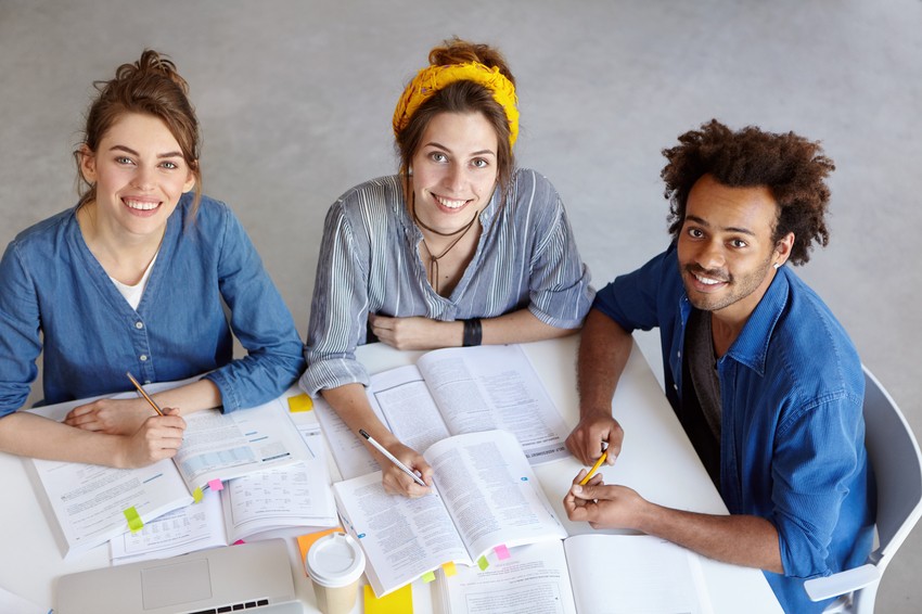 Three people sitting at a table, smiling and working with open textbooks and notebooks, balancing health and study as they prepare for the MCCQE1 exam. They have various items like highlighters, a coffee cup, and Ace QBank resources spread out on the table.