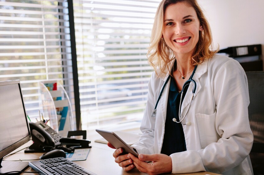 A smiling doctor in a white coat and stethoscope sits at a desk with a tablet in her hands, a computer displaying Ace QBank and office supplies in the background, ideal for MCCQE1 exam preparation.