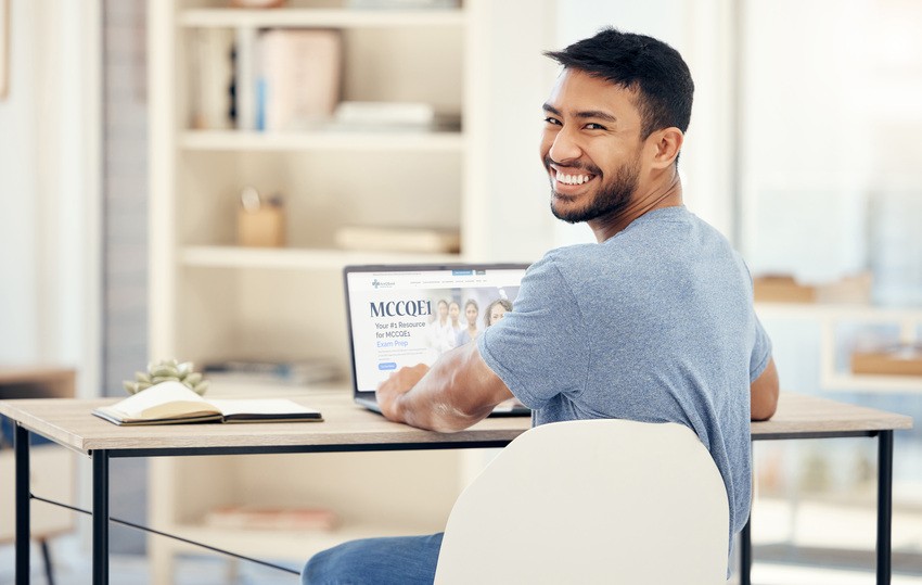 A man sits at a desk, smiling at the camera while using a laptop. An open notebook and a pen are on the desk, as he delves into MCCQE1 exam preparation. Shelves with books and decor items are visible in the background.