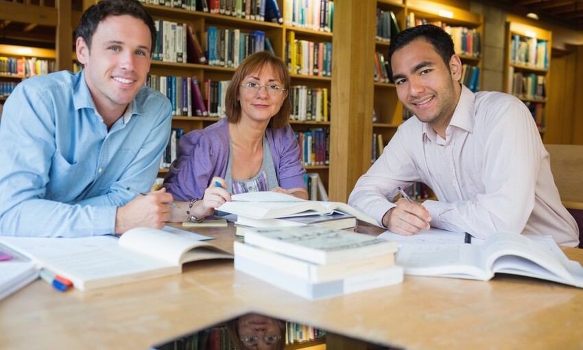 Three people sit at a table in a library with open books, smiling at the camera. Shelves filled with books are in the background, with one prominently holding an "Acing MCCQE1" guide – a testament to their dedication to unlocking their future success in Canada.