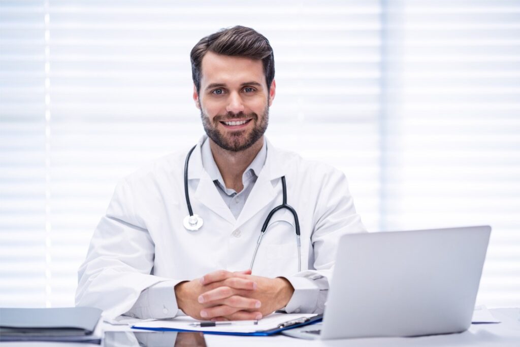 A male doctor in a white coat with a stethoscope around his neck sits at a desk with a laptop, smiling, in front of a window with blinds, as he reviews materials on Ace QBank for his MCCQE1 Exam Preparation.