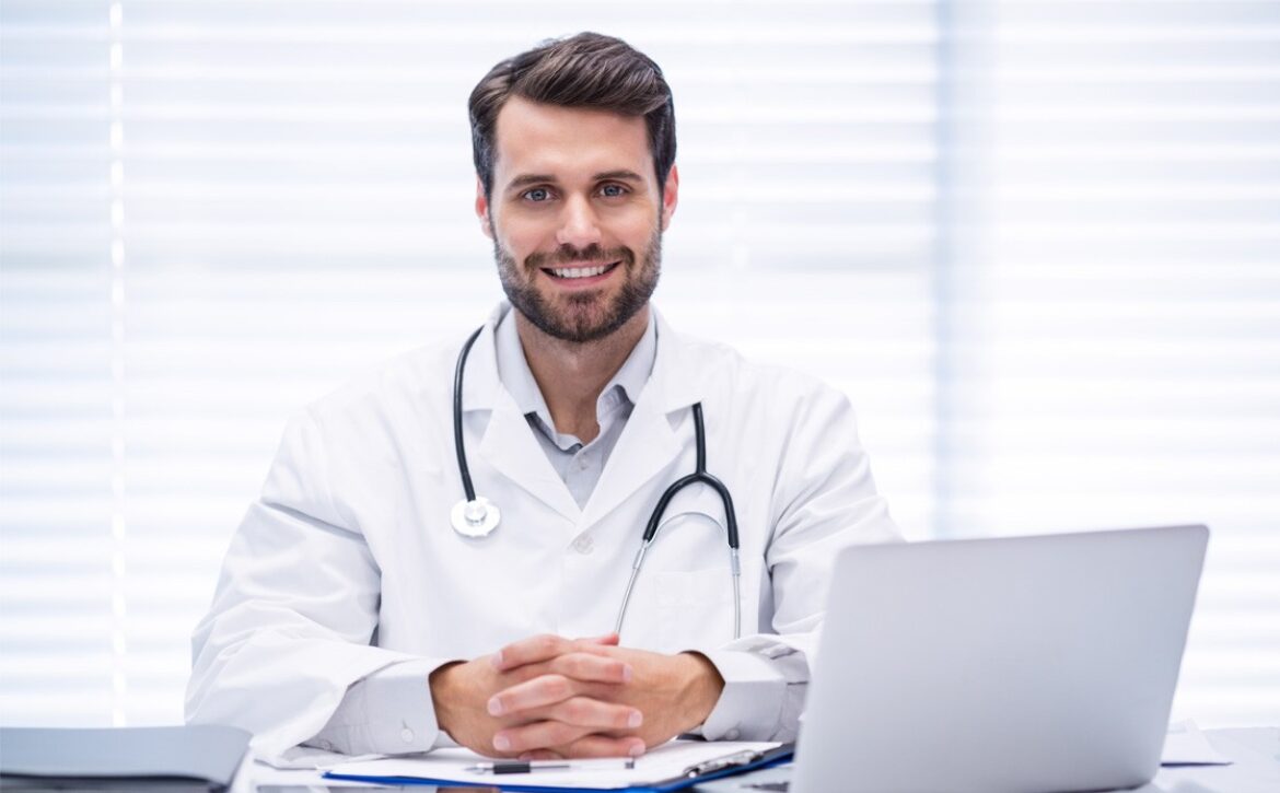 A male doctor in a white coat with a stethoscope around his neck sits at a desk with a laptop, smiling, in front of a window with blinds, as he reviews materials on Ace QBank for his MCCQE1 Exam Preparation.